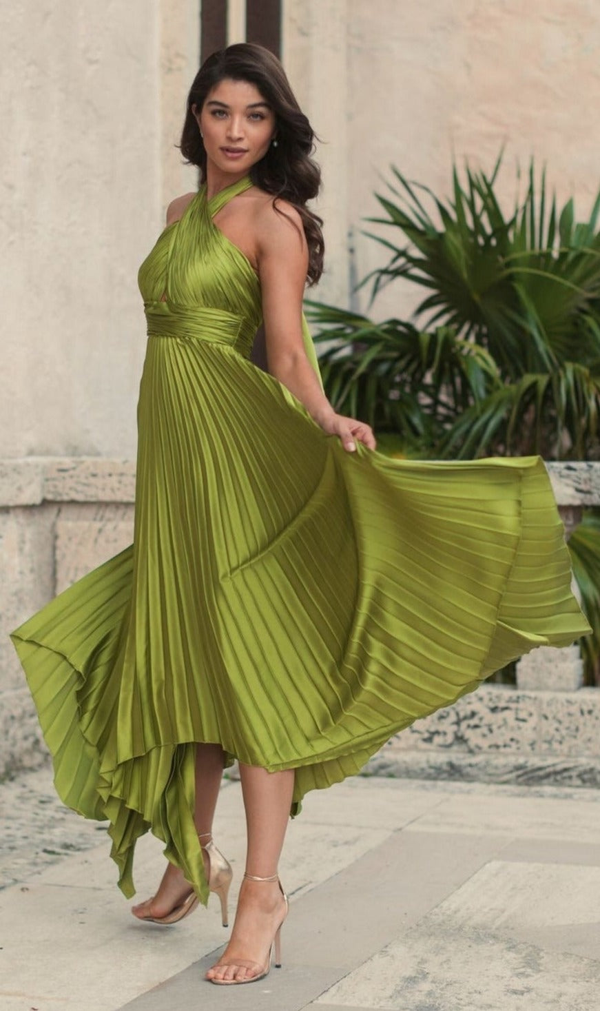 A woman poses outdoors in the Long Formal Dress 11302 by Mac Duggal, an olive green pleated halter gown with an asymmetrical skirt and nude heels, gracefully holding out one side of the dress.